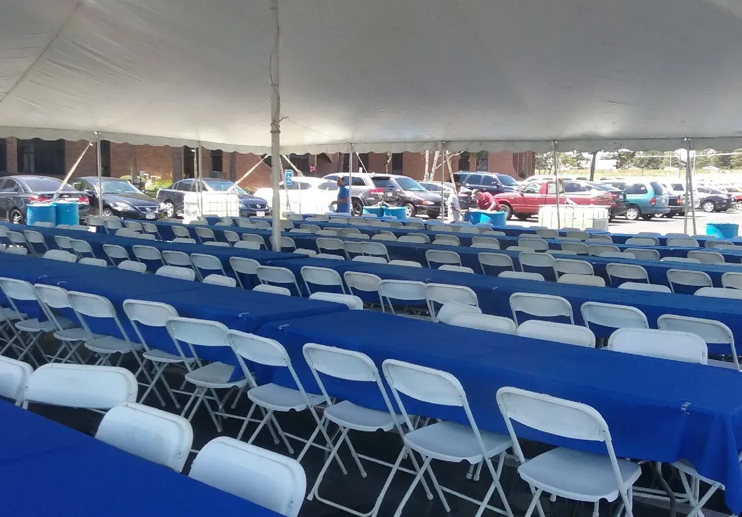 Long rows of tables with royal blue tablecloths and white folding chairs under a white tent, with a parking lot of cars beyond