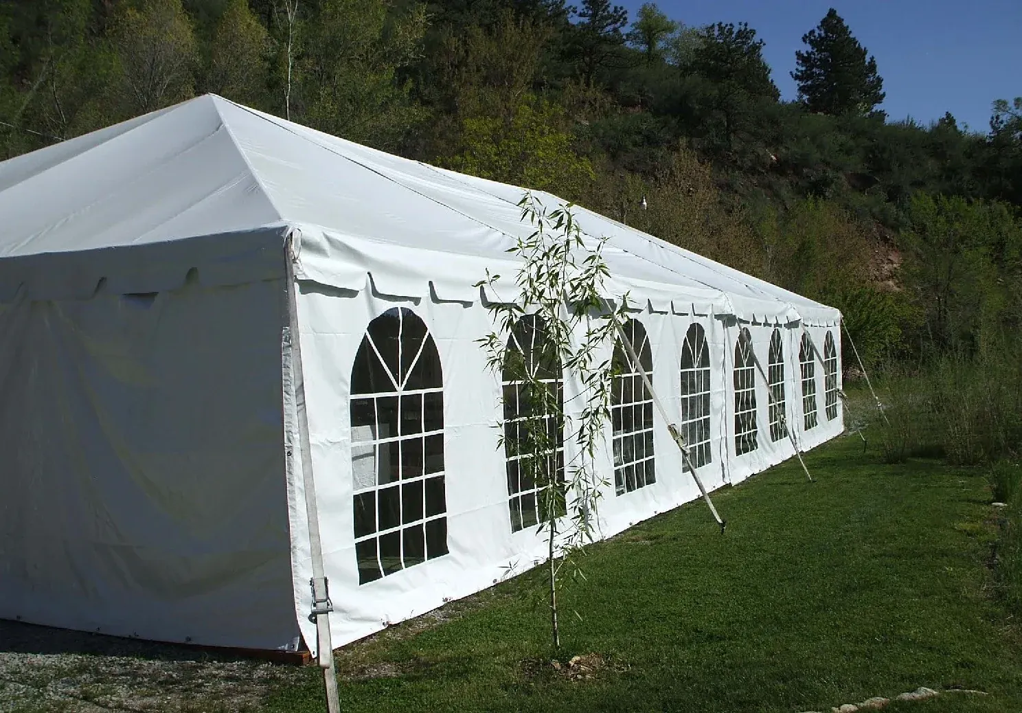 Long white gable tent with arched, gridded windows along its sidewall on a green lawn below a wooded hillside