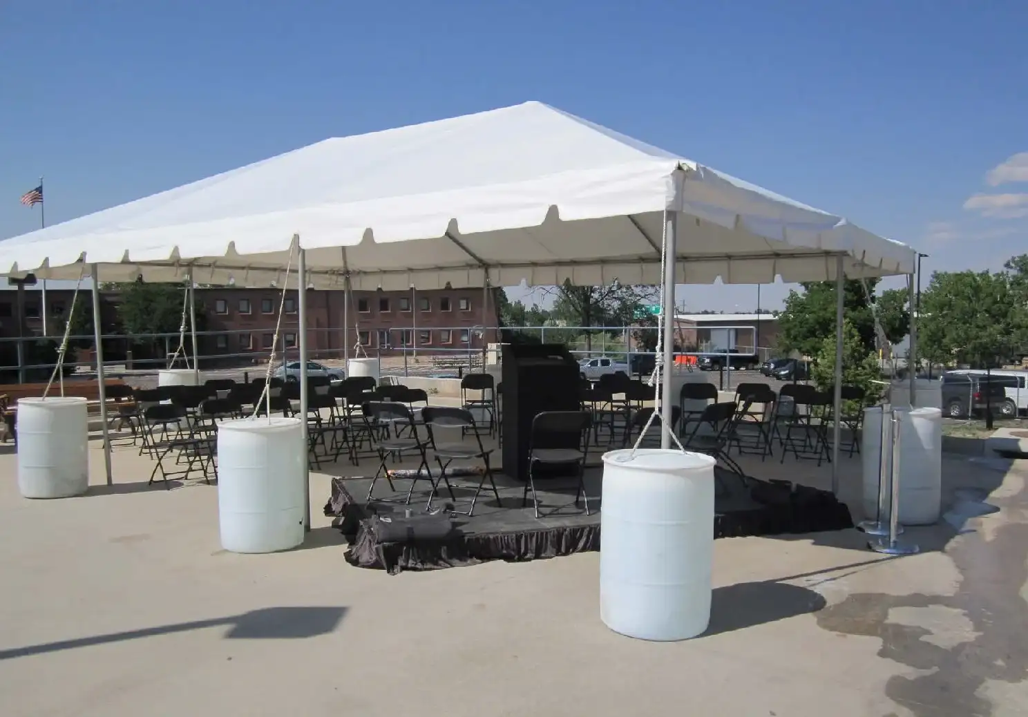 White frame tent over rows of black folding chairs and a black-skirted stage with a podium, white barrels at the legs, on a paved lot