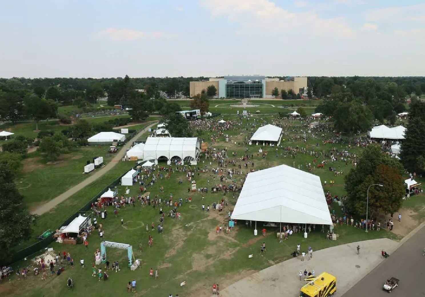 Aerial view of a crowded park event with a large white clearspan tent, smaller white tents and a yellow truck beside the road