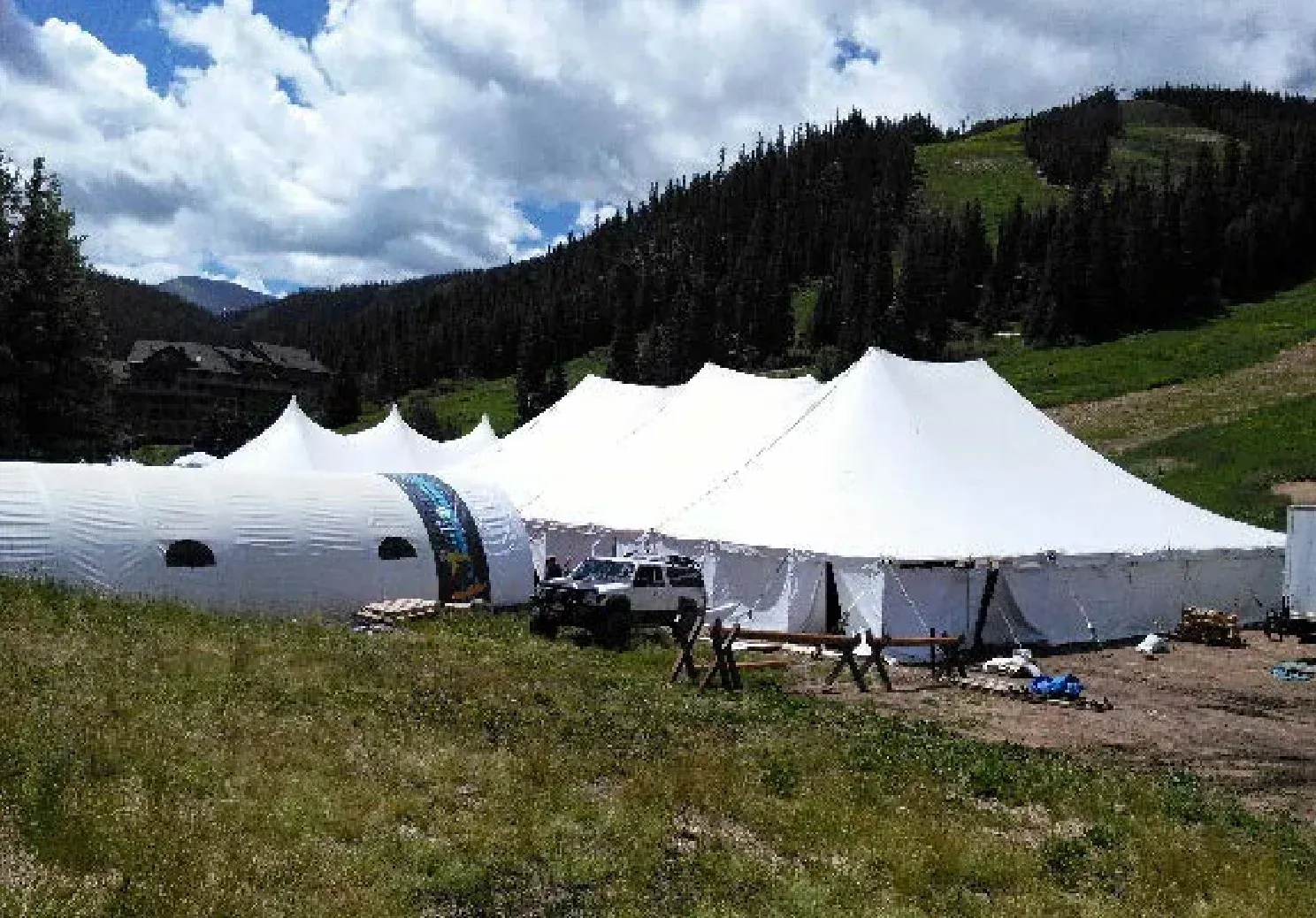 White pole tents beside an arched white tent with oval windows on a mountain meadow, with an SUV in front and evergreen slopes behind