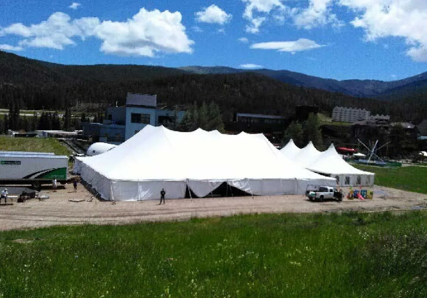 Large white pole tent with several peaks and smaller tents on a dirt lot, with a trailer, a pickup and mountain slopes behind