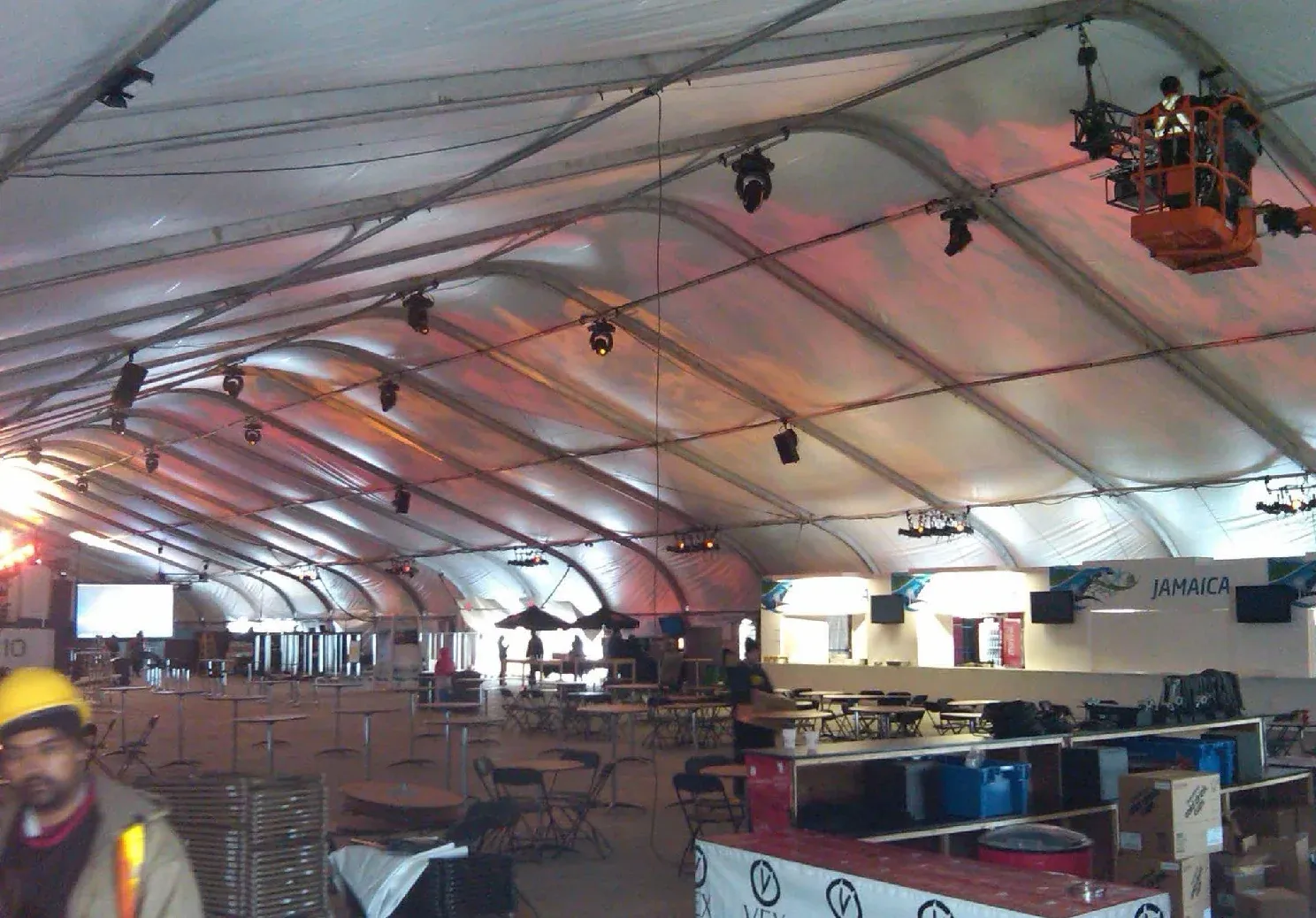 Inside a large clearspan tent during setup, with stage lights overhead, an aerial lift, cocktail tables and a worker in a hard hat