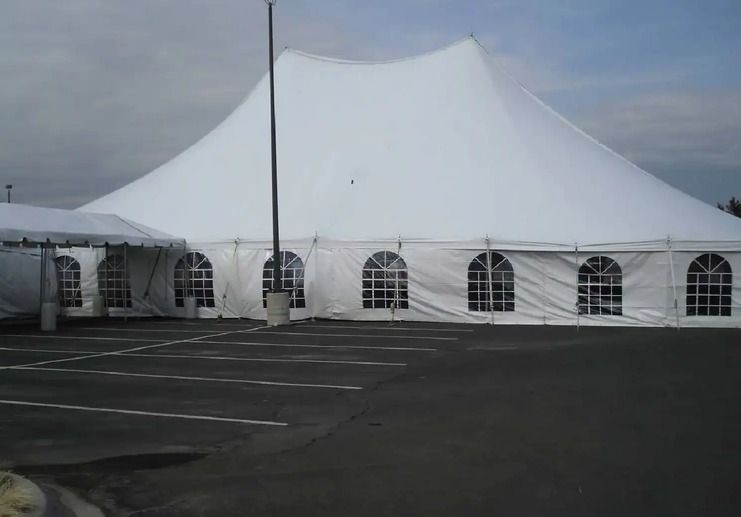 Large white pole tent with arched window sidewalls and a smaller frame tent at left, in an asphalt parking lot under an overcast sky
