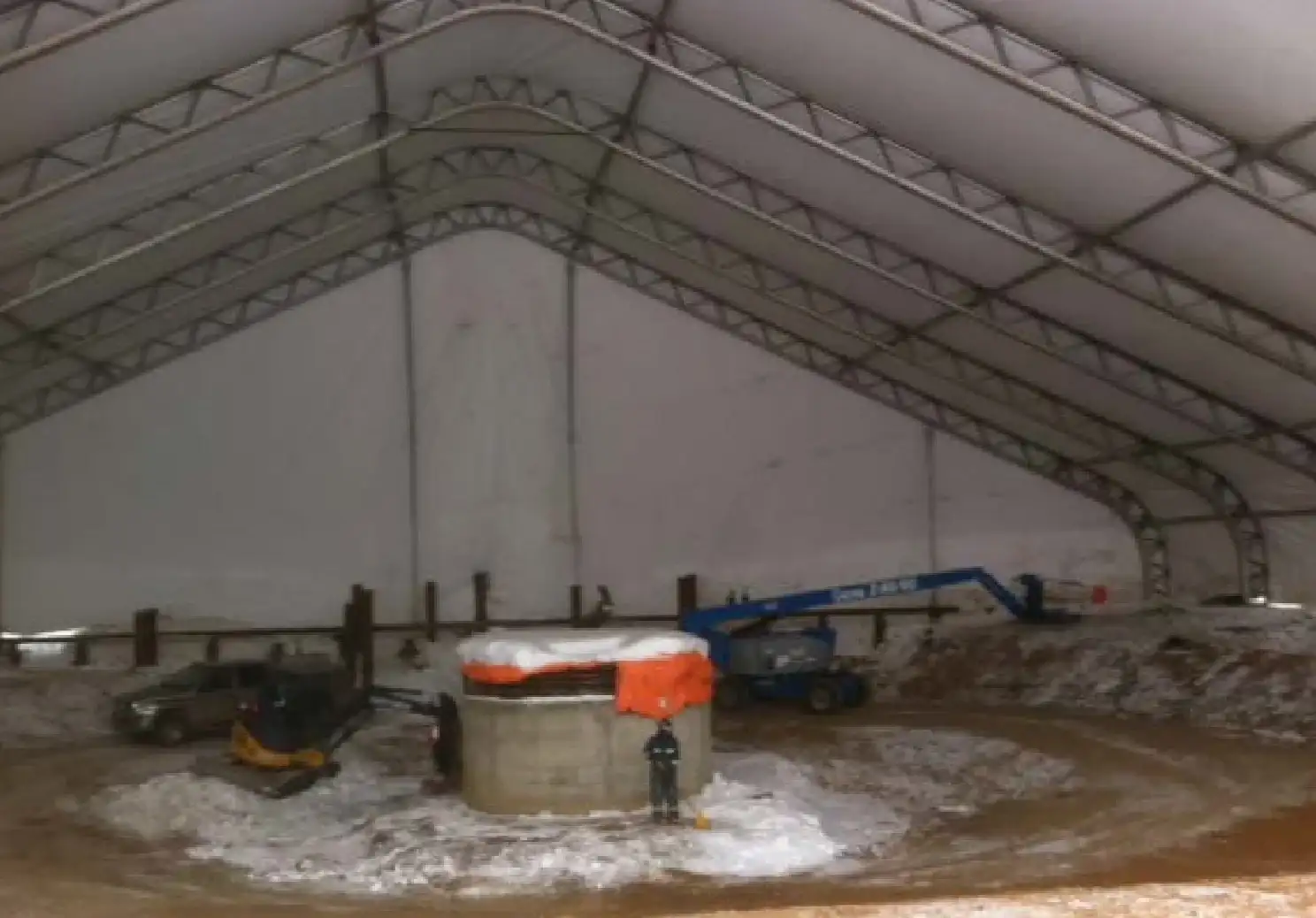 Inside a truss-frame clearspan tent over a snowy dirt floor, with a boom lift, mini excavator and a round concrete structure