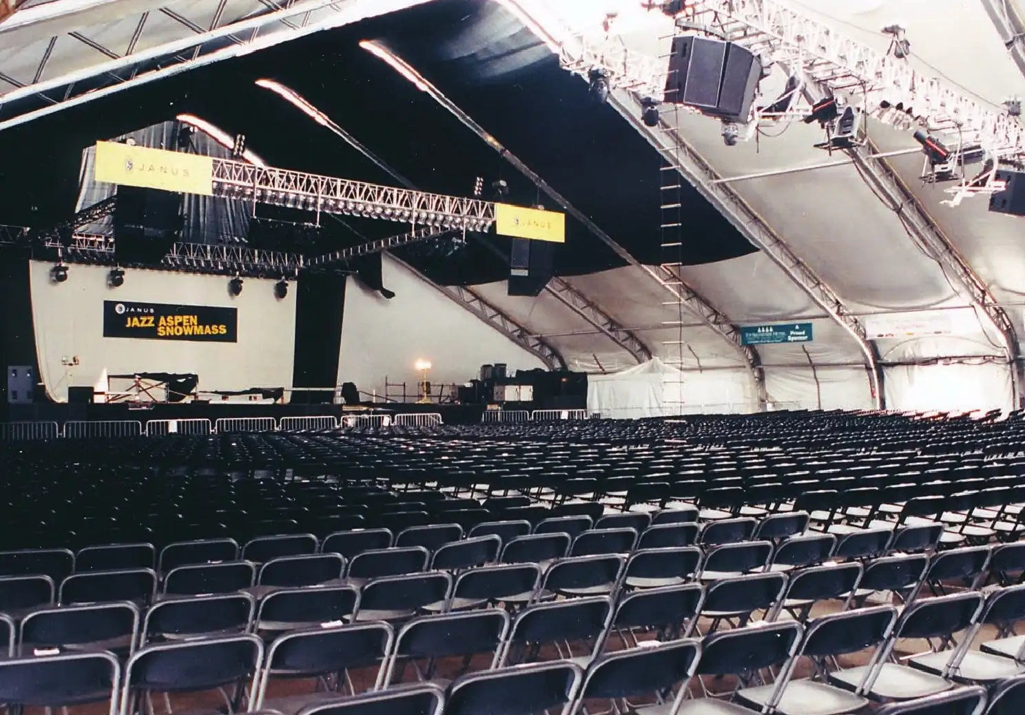 Rows of black folding chairs inside a large clearspan tent facing a stage with truss rigging, speakers and hanging banners