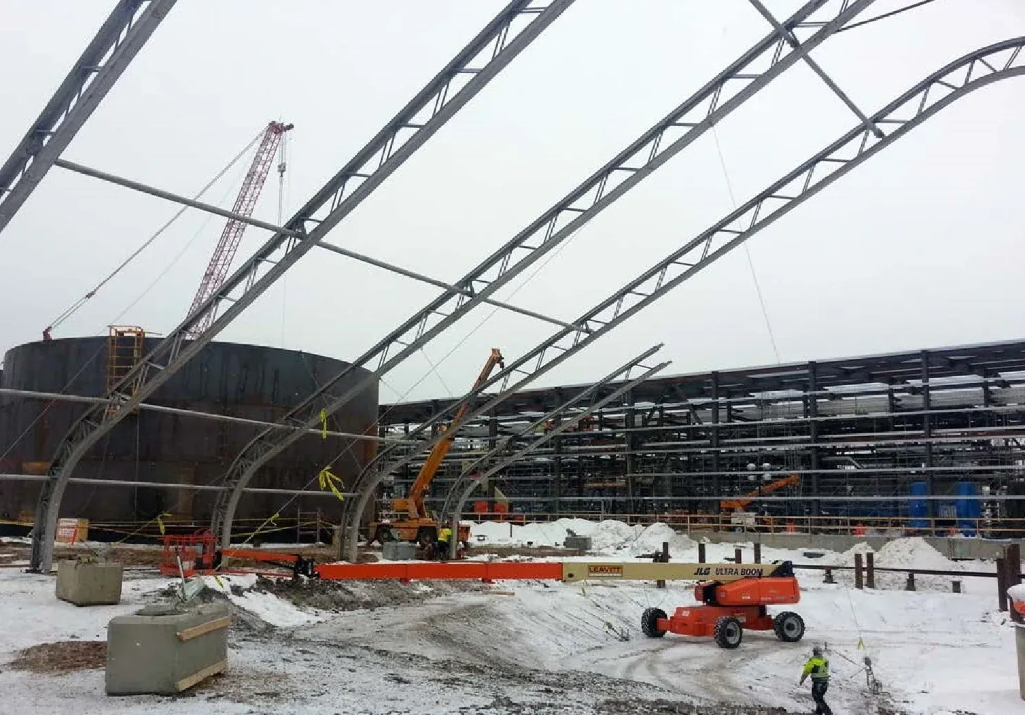 Steel truss arches of a clearspan tent frame going up at a snowy industrial site, with a boom lift, cranes and a large dark tank behind