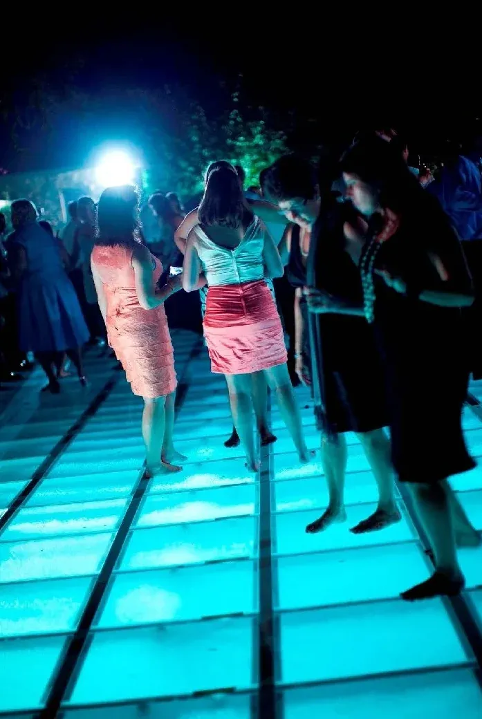 Guests standing on a clear-panel floor lit cyan from below at a night party