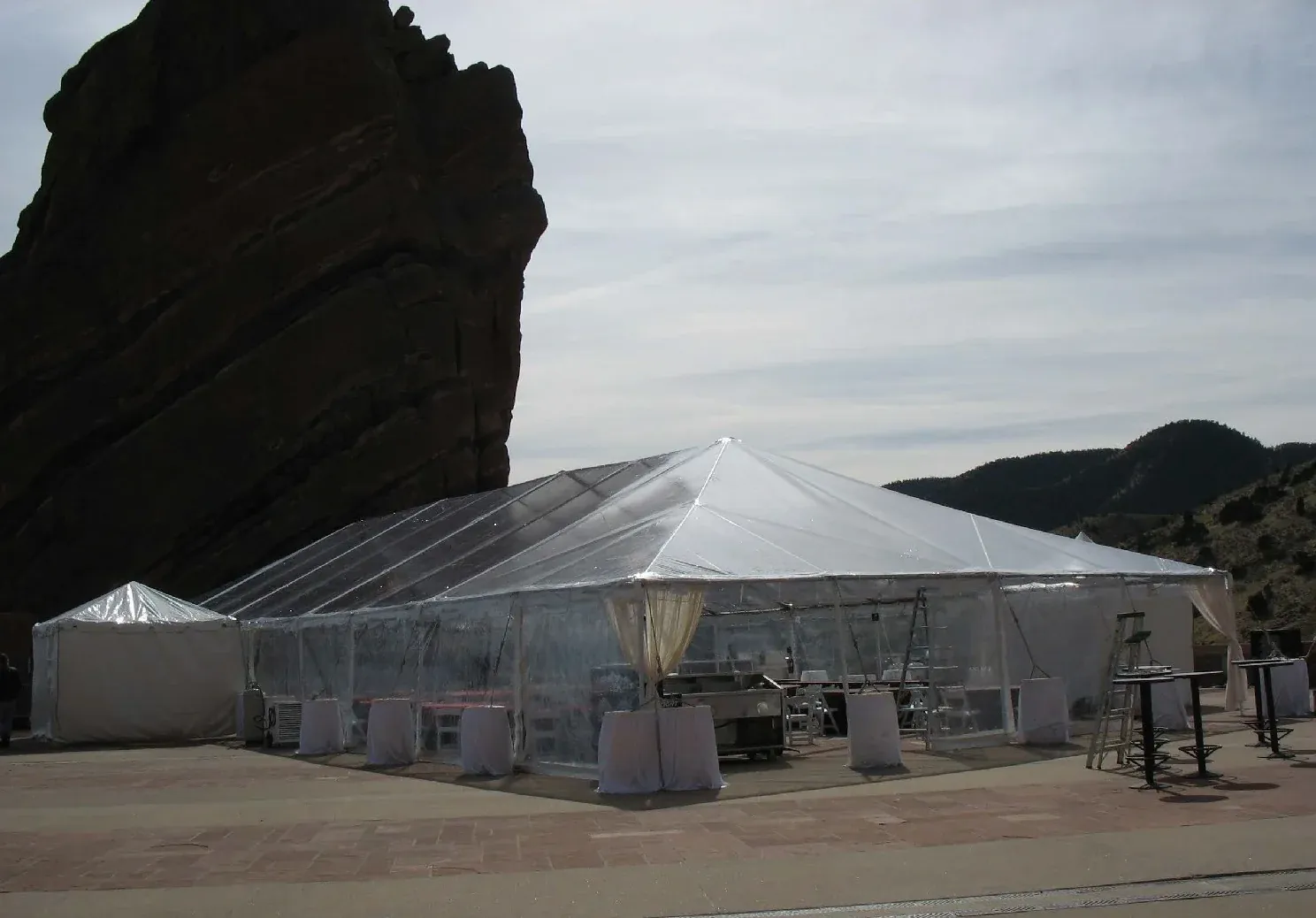 Clear-top frame tent with clear sidewalls, gathered drapes and linen-covered high tables on a paved terrace beside a dark rock face