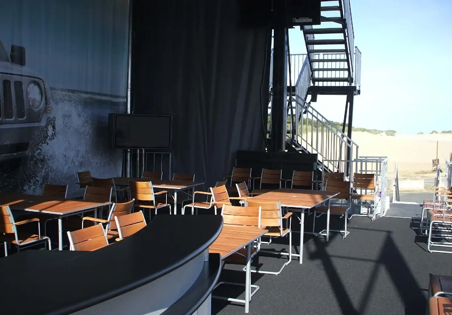 Wood-top tables and slatted chairs on black carpet in front of black drapes, with a curved bar, a TV and a steel staircase at right
