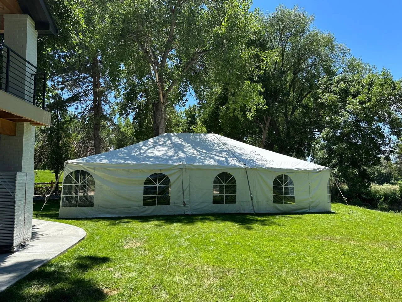 White frame tent with arched window sidewalls on a green lawn under tall trees